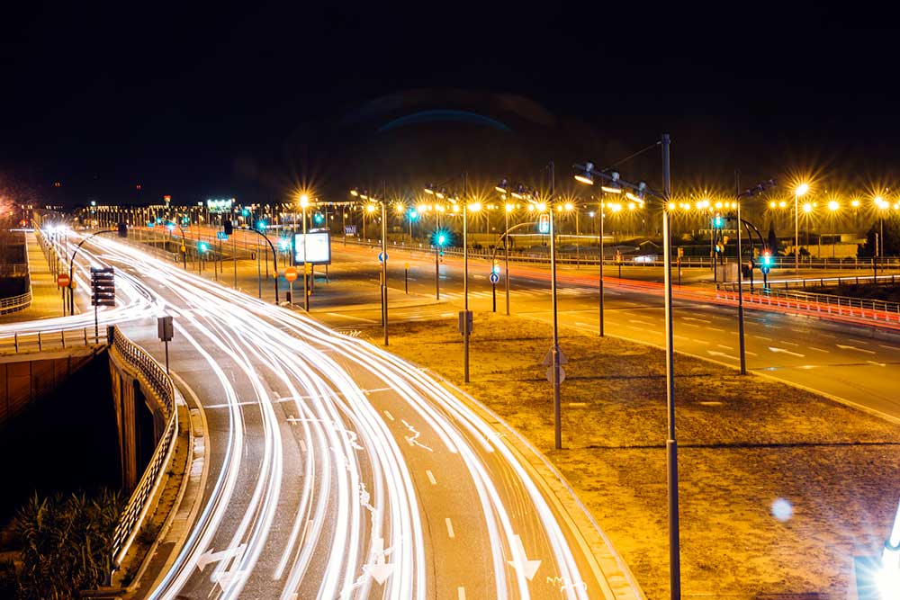 A vibrant night scene of a city street with bright streetlights and long exposure light trails from passing vehicles, showcasing urban life and motion.