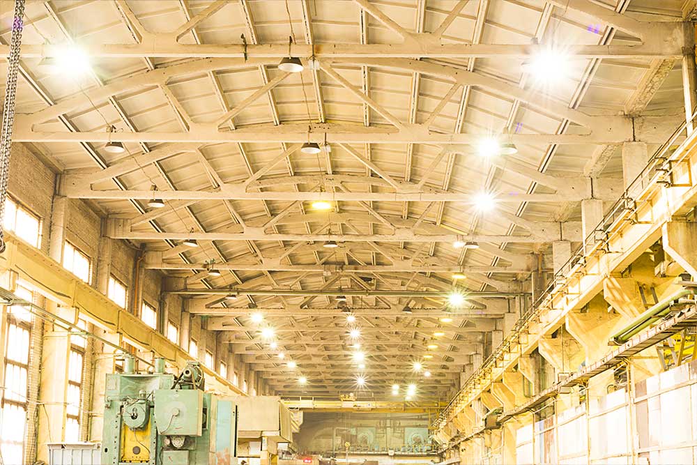 Brightly lit ceiling of a manufacturing factory, showcasing structural beams, industrial lighting, and a spacious production environment.