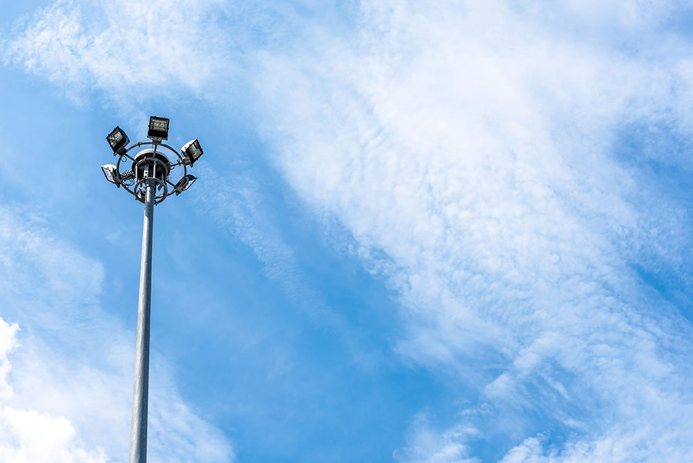 A high electric light post with multiple spotlights pointing in various directions, set against a bright blue sky with scattered clouds.